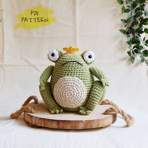 Op de afbeelding: Een groene gehaakte kikker met een gele kroon zit op een ronde houten basis. De kikker heeft grote, witte ogen en een kleine, zwarte mond. De basis is omgeven door een bruin touw.