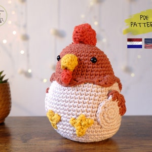 Op de afbeelding: Een gehaakt amigurumi-speelgoed in de vorm van een kip met een rode kam, gele snavel en zwarte ogen. De kip zit op een houten oppervlak. De afbeelding bevat ook de tekst "PDF PATTERN" en vlaggen van Nederland en de Verenigde Staten.