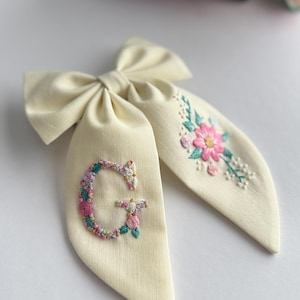 Peut inclure: Nœud papillon crème avec broderie florale. Le nœud présente un grand nœud décoratif et deux longues queues. L'une des queues affiche la lettre "G" brodée entourée de fleurs. L'autre queue a un motif floral. L'arrière-plan est flou.