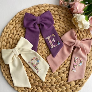 May include: Three fabric hair bows with embroidered floral letters. The bows are in purple, pink, and cream colors. The letters are E, P, and P.