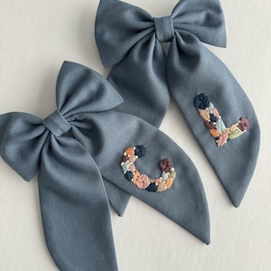 May include: Two blue fabric hair bows with embroidered floral letters, one with a "C" and the other with an "L".