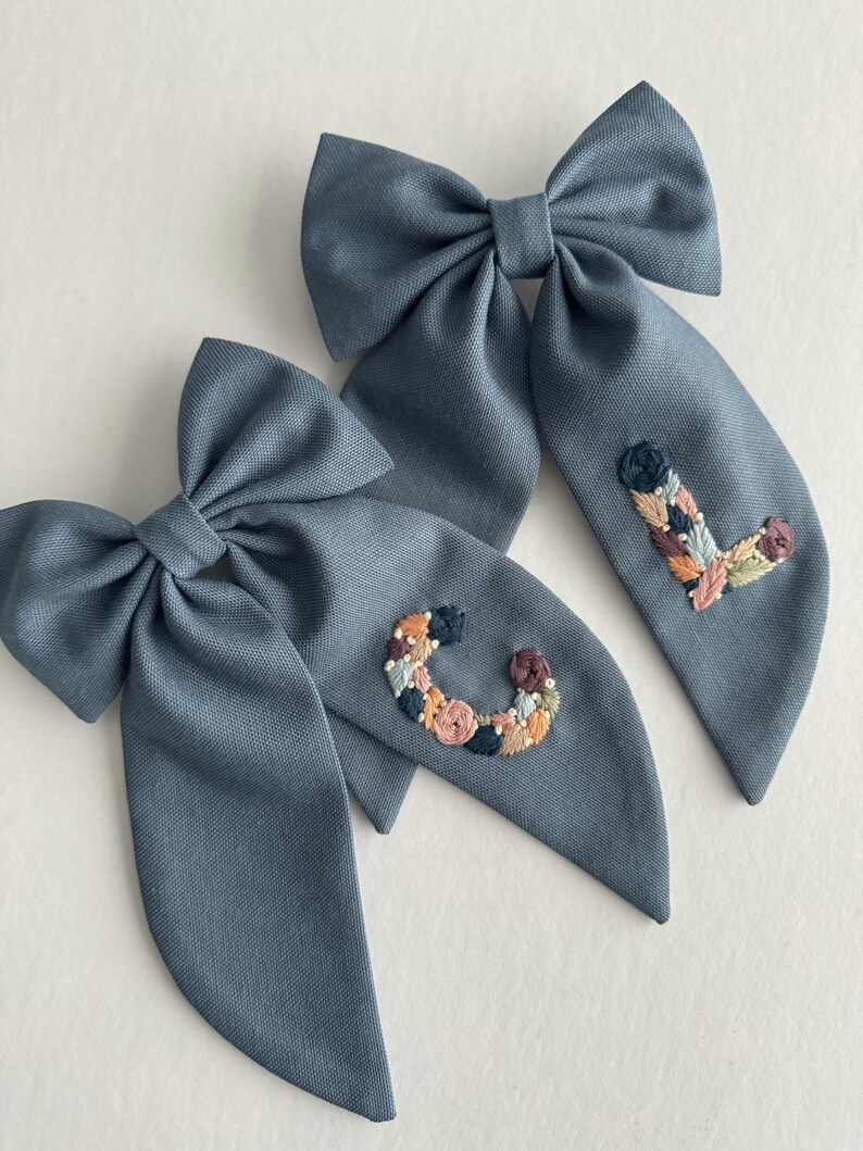 May include: Two blue fabric hair bows with embroidered floral letters, one with a "C" and the other with an "L".