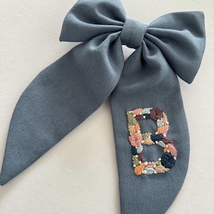 May include: A blue fabric hair bow with a large, embroidered floral letter "B" in the center.