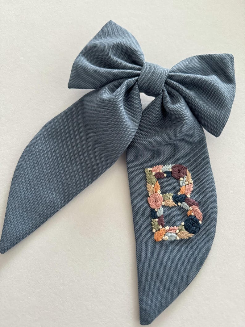 May include: A blue fabric hair bow with a large, embroidered floral letter "B" in the center.