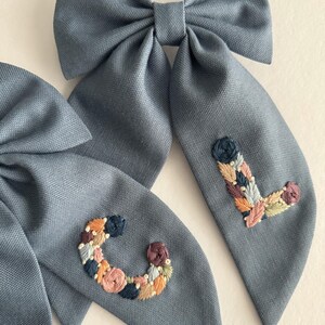 May include: Two blue fabric hair bows with embroidered floral letters, one with a "C" and the other with an "L".