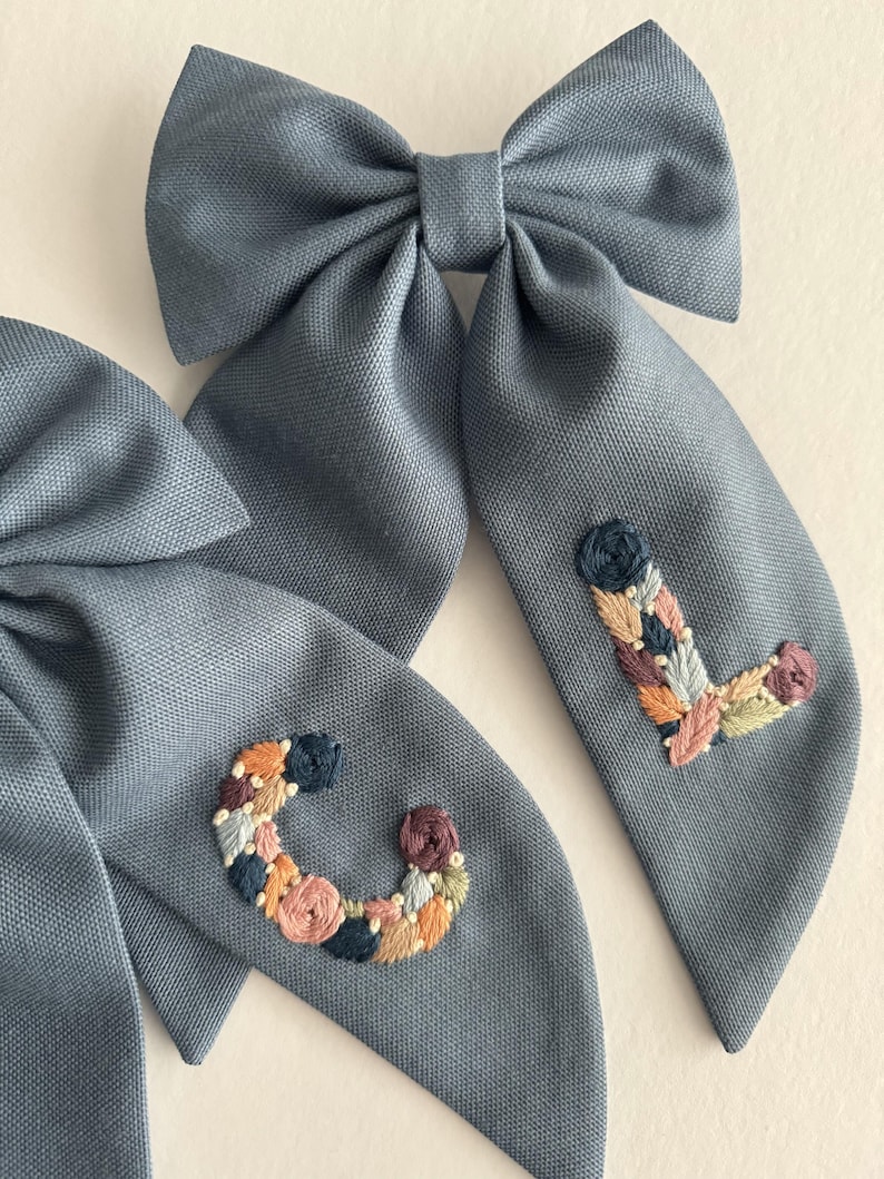 May include: Two blue fabric hair bows with embroidered floral letters, one with a "C" and the other with an "L".