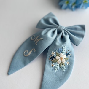 Pode incluir: Um laço de cabelo azul claro com detalhes florais bordados e as iniciais "MS". O laço apresenta uma forma de laço grande com caudas longas e fluidas. O bordado floral inclui detalhes em branco, azul e verde.