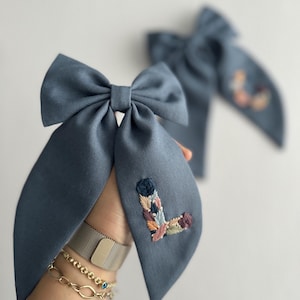 May include: A blue fabric bow with a colorful embroidered letter L. The bow is tied with a knot in the center and has two long tails.
