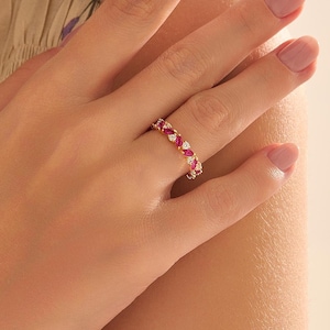 May include: A gold band ring featuring an alternating pattern of marquise-cut red gemstones and round-cut clear white stones. The ring is worn on a finger, showcasing the continuous arrangement of the colorful stones set in a polished gold metal.
