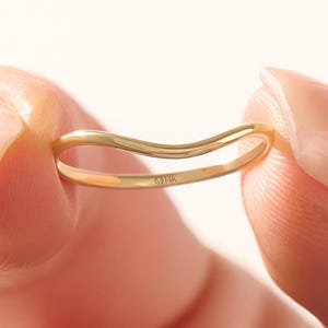May include: A delicate, curved gold ring held between two fingers. The ring has a smooth, polished finish and a subtle wave design. The ring is a simple, elegant piece of jewelry.