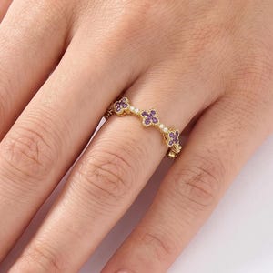 May include: A gold ring featuring a series of small, four-petaled flower motifs. Each flower is centered with a tiny white pearl and accented with small purple stones on the petals. The band is thin and metallic, encircling the finger.