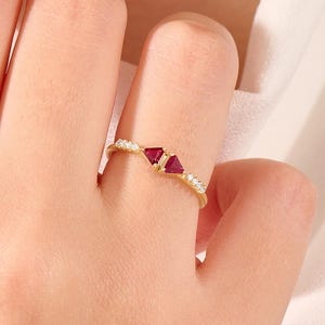 May include: A gold ring featuring two triangular red gemstones and small white gemstones. The ring is set on a delicate gold band. The ring is shown on a person's finger.