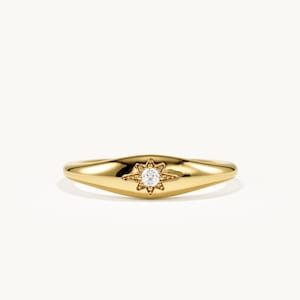 May include: A polished gold band ring featuring a central star-shaped setting. The star motif is engraved with fine textured details and holds a single, small, clear round gemstone at its center, creating a bright focal point on the metal.