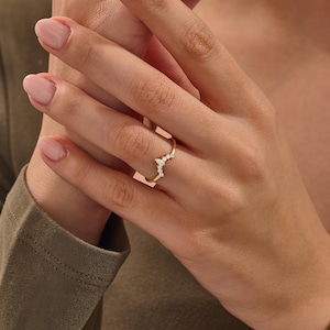 Pode incluir: Um anel dourado com um design curvo, cravejado com pequenas pedras preciosas transparentes. O anel é usado em um dedo, destacando sua aparência delicada e elegante. O design do anel é uma versão moderna de um estilo clássico.