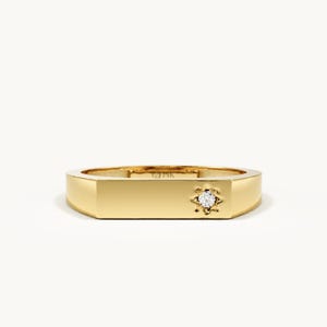 May include: A polished gold band ring featuring a flat, rectangular top surface. A single, small, clear round gemstone is set within a star-shaped engraving on the right side of the flat surface. The interior of the band is marked with the letters MK.