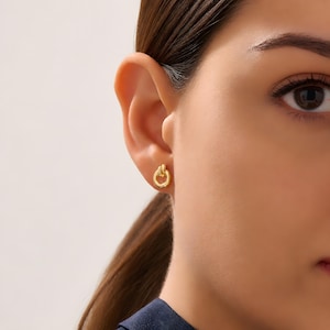 K&ouml;nnte beinhalten: Ein kleiner goldener Ohrstecker mit einem polierten, kreisf&ouml;rmigen Ringdesign und einem zarten Knotendetail an der Oberseite. Der Ohrstecker ist aus glattem, reflektierendem goldfarbenem Metall gefertigt und wird mit einem Stift befestigt.