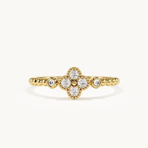 May include: A gold ring featuring a central four-petaled flower motif composed of four round clear stones and a small central gold bead. The band is crafted with a beaded texture and includes two additional round clear stones on each side.