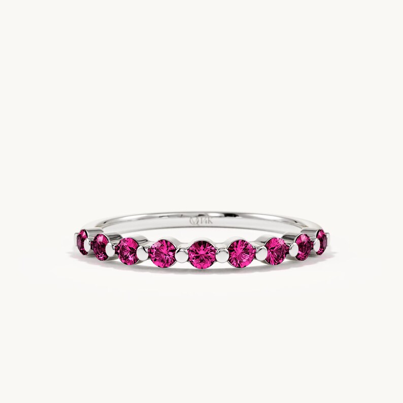 K&ouml;nnte beinhalten: Ein silberfarbener Ring mit einer Reihe von neun rund geschliffenen, leuchtend pinkfarbenen Edelsteinen in einer gemeinsamen Krappenfassung. Die Innenseite des Rings ist deutlich mit 14K graviert, was den Reinheitsgrad des Metalls angibt.