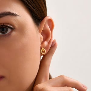 K&ouml;nnte beinhalten: Ein kleiner goldfarbener Ohrstecker mit einem kreisf&ouml;rmigen Ringdesign und einem geknoteten Detail an der Oberseite. Der Ohrring hat eine polierte metallische Oberfl&auml;che und ist am Ohrl&auml;ppchen befestigt, was eine einfache und kompakte Struktur zeigt.