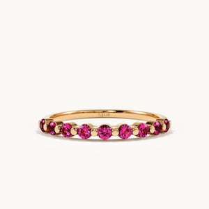 K&ouml;nnte beinhalten: Ein Goldring mit einer Reihe von neun rund geschliffenen, leuchtend pinkfarbenen Edelsteinen in einer gemeinsamen Krappenfassung. Auf der Innenseite des Rings ist deutlich die Gravur 14K zu sehen, die den Goldgehalt des Schmuckst&uuml;cks angibt.