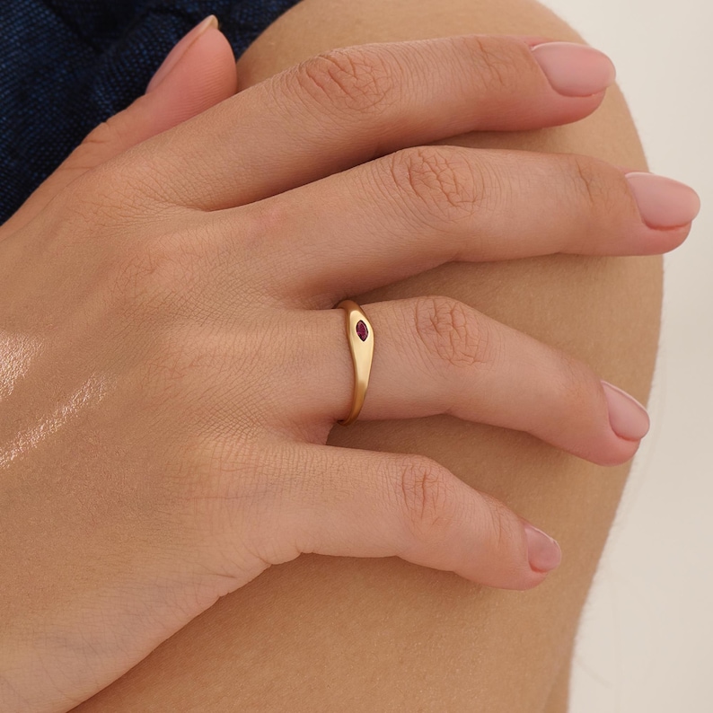 May include: A gold ring with a small, oval-shaped ruby gemstone. The ring has a simple, elegant design and is worn on a finger. The ring is a piece of jewelry.