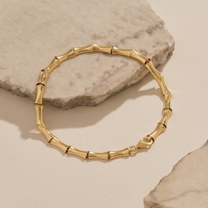 Op de afbeelding: Een goudkleurige armband bestaande uit herhalende, gepolijste, cilindrische segmenten die lijken op bamboestengels. De armband is cirkelvormig en is aan één uiteinde voorzien van een klein, veilig karabijnsluitingsmechanisme.