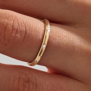 May include: A gold ring with three small, clear gemstones set into the band. The ring has a smooth, polished finish and is shown on a finger. The ring is a simple, elegant piece of jewelry.