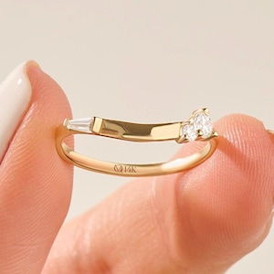 May include: A 14K gold ring featuring a curved band with a single baguette-cut diamond on one side and two round brilliant-cut diamonds on the other. The band is engraved with the text 14K on the inner surface, showcasing a polished finish.