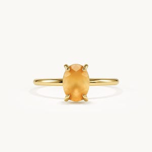 May include: A gold ring featuring a central oval-shaped orange gemstone held in place by four prongs. The band is thin, smooth, and polished, creating a continuous circular shape that supports the faceted stone in a vertical orientation.