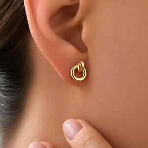 K&ouml;nnte beinhalten: Ein kleiner goldener Ohrstecker mit kreisf&ouml;rmigem Ringdesign und polierter Oberfl&auml;che. Der Ohrstecker besteht aus einem glatten, durchgehenden Goldring, der an einer kleineren, vertikalen Goldschlaufe befestigt ist und eine geometrische Form bildet.