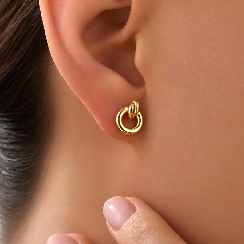 K&ouml;nnte beinhalten: Ein kleiner goldener Ohrstecker mit kreisf&ouml;rmigem Ringdesign und polierter Oberfl&auml;che. Der Ohrstecker besteht aus einem glatten, durchgehenden Goldring, der an einer kleineren, vertikalen Goldschlaufe befestigt ist und eine geometrische Form bildet.