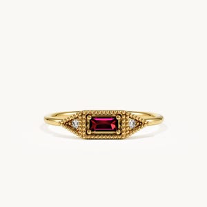 May include: A gold ring featuring a central rectangular baguette-cut deep red garnet stone, flanked by two small triangular clear stones, all set within a beaded metal frame on a smooth, polished gold band.