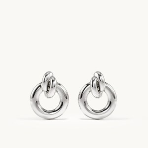 K&ouml;nnte beinhalten: Ein Paar Ohrringe in polierter Silberoptik mit einem kreisf&ouml;rmigen Ringdesign, das an einem kleineren, geknoteten Schlaufenelement h&auml;ngt. Die Ohrringe haben eine glatte, reflektierende Metalloberfl&auml;che und eine klare, symmetrische Struktur ohne Verzierungen.