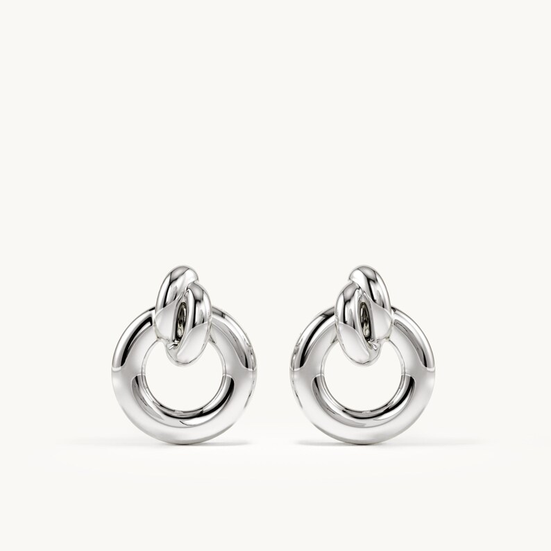K&ouml;nnte beinhalten: Ein Paar Ohrringe in polierter Silberoptik mit einem kreisf&ouml;rmigen Ringdesign, das an einem kleineren, geknoteten Schlaufenelement h&auml;ngt. Die Ohrringe haben eine glatte, reflektierende Metalloberfl&auml;che und eine klare, symmetrische Struktur ohne Verzierungen.