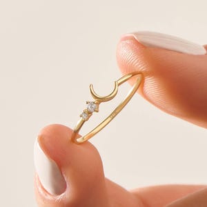 Puede incluir: Un delicado anillo de oro con un diseño de media luna y dos pequeñas gemas brillantes. El anillo se sostiene entre los dedos, mostrando sus finos detalles y su elegante simplicidad. Es una pieza de joyería.