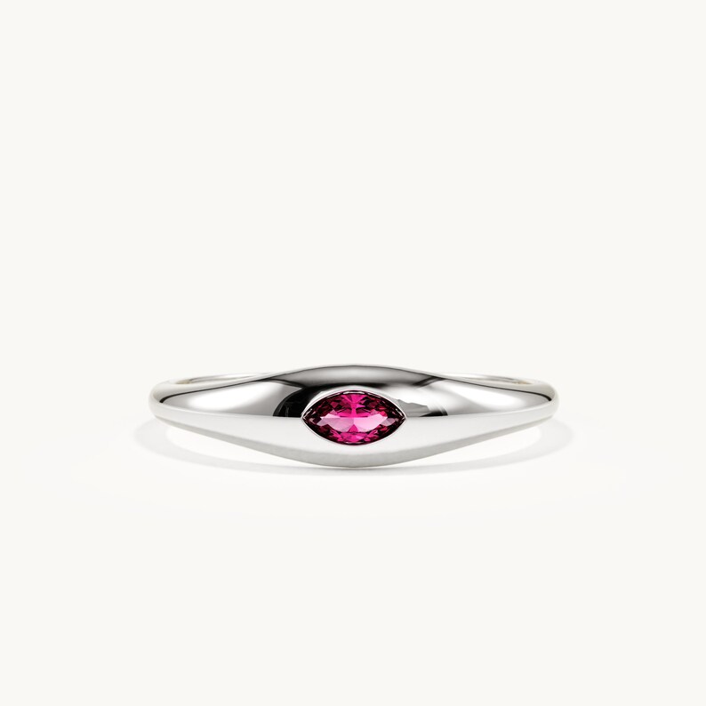 May include: A polished silver-tone ring featuring a smooth, tapered band and a single marquise-cut vibrant pink gemstone set horizontally in the centre. The metal surface has a high-shine finish that reflects light across the curved band.