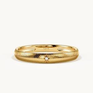 May include: A yellow gold band ring featuring a textured herringbone pattern across the outer surface. A single small round white diamond is set in the center of the band within a star-shaped engraving. The interior of the ring is stamped with 18K.