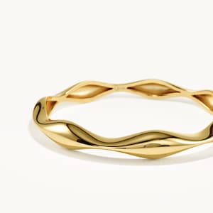 May include: A polished gold ring featuring a continuous wavy, undulating band design. The metal surface is smooth and reflective, catching the light along its curved, organic shape. The ring has a consistent yellow gold tone throughout its structure.