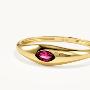 May include: A 14K gold ring featuring a smooth, polished band with a central marquise-cut vibrant pink gemstone set flush into the metal. The interior of the band is clearly stamped with the text 14K, indicating the gold purity of the jewelry piece.