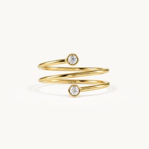 Pode incluir: Um anel de ouro com design em espiral, apresentando duas pedras redondas, transparentes e facetadas, fixadas em engastes de bisel em cada extremidade. A banda é de ouro amarelo polido e tem uma estrutura de bobina aberta que envolve o dedo.