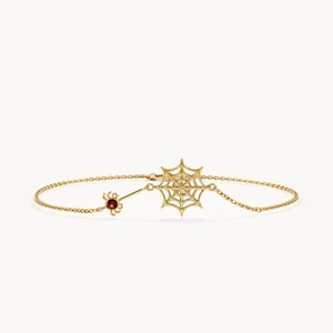 May include: A gold chain bracelet featuring a central spider web charm and a small spider accent. The spider has a round, deep red gemstone set in its body. The bracelet is constructed from a delicate gold link chain with a polished finish.