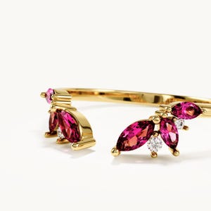 May include: A gold open ring featuring two clusters of marquise-cut vibrant pink gemstones and small round clear stones. The band is polished gold, and the design is symmetrical, with the pink stones arranged in a leaf-like pattern on both ends.