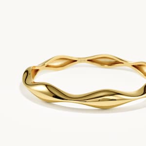 May include: A polished gold bangle bracelet featuring a continuous, undulating wave-like design. The metal surface is smooth and reflective, creating a series of rounded, organic curves that form the circular shape of the jewellery piece.
