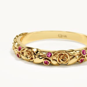 May include: A 14K gold ring featuring a sculpted band with a repeating pattern of polished gold roses and leaves. The design is accented with small, round, vibrant pink gemstones set within the metalwork. The interior is stamped with 14K.