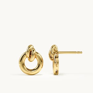 May include: A pair of polished gold stud earrings featuring a circular hoop design with a smaller, interlocking knot-like loop at the top. The earrings have a smooth, reflective metallic finish and include a standard post backing for secure attachment.