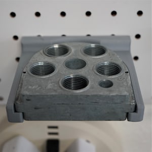 May include: A metallic gray industrial component with multiple threaded holes of varying sizes. The component is mounted on a gray bracket, and the background is a white pegboard with black holes.