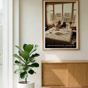 May include: A framed poster featuring John Lennon and Yoko Ono in bed, with signs reading "Hair Peace" and "Bed Peace." The poster is in a light wood frame, hanging above a wooden cabinet. A potted plant sits to the left of the cabinet.