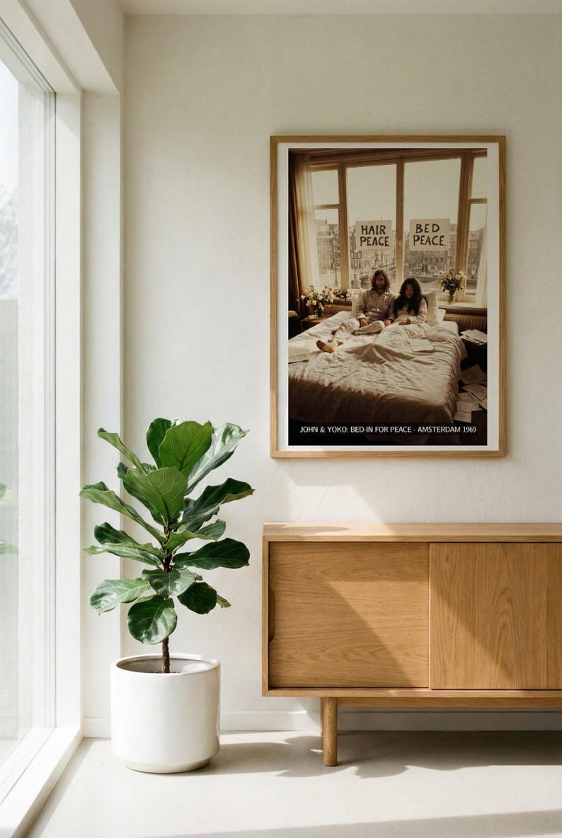 May include: A framed poster featuring John Lennon and Yoko Ono in bed, with signs reading "Hair Peace" and "Bed Peace." The poster is in a light wood frame, hanging above a wooden cabinet. A potted plant sits to the left of the cabinet.