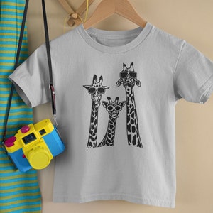 May include: A gray t-shirt with a graphic of three giraffes wearing sunglasses. The giraffes are black and white and are standing in a row. The t-shirt is on a hanger.
