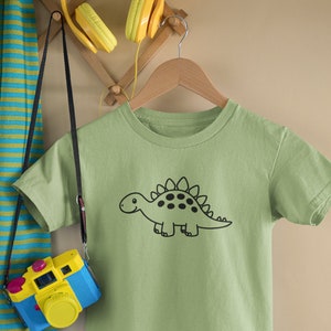 May include: A light green t-shirt with a black and white cartoon dinosaur graphic. The dinosaur is facing right and has a smiling face. The t-shirt is hanging on a wooden hanger.